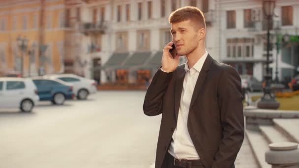商人在城市广场积极通电话 背景是日落城市景观 穿着黑色套房的年轻人在街上用手势在手机上说话 温暖的晨光 — 图库视频影像