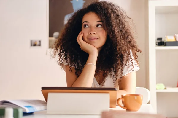 Joven hermosa mujer con el pelo rizado oscuro sentado en la mesa con la tableta y la taza de café cerca de apoyarse en la mano soñadoramente mirando a un lado pasar tiempo en casa —  Fotos de Stock