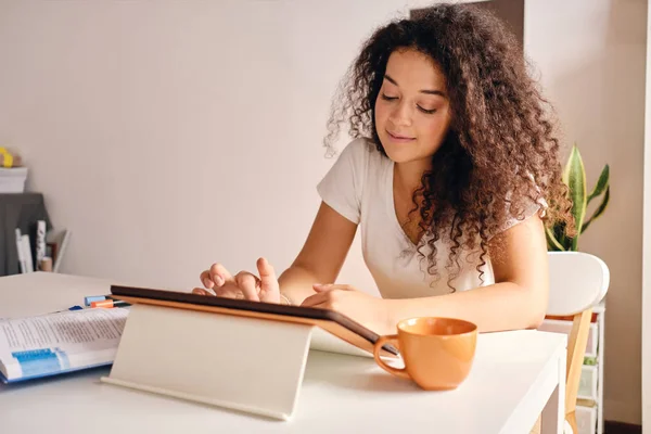 漂亮的微笑的女孩与黑卷发坐在桌子与书和咖啡杯梦想使用平板电脑舒适的家 — 图库照片