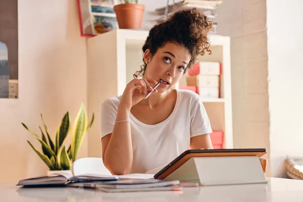 Chica bonita con el pelo rizado oscuro sentado en la mesa con la tableta soñando mirando hacia arriba con lápiz en la mano estudiando en la casa acogedora moderna —  Fotos de Stock