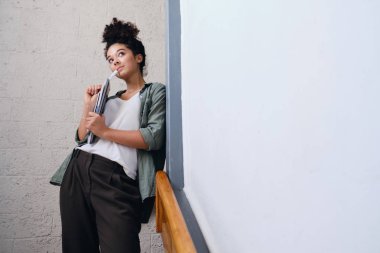 Haki gömlek ve pantolon elinde ders kitapları ile duvara yaslanmış koyu kıvırcık saçlı genç güzel öğrenci kız elinde ve hayali bir kenara bakıyor