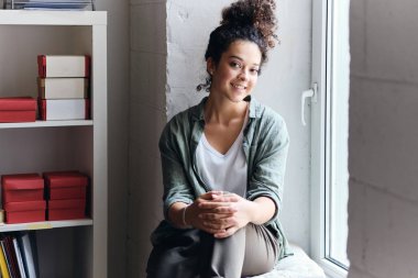 Üniversite seyircisi pencere eşik üzerinde oturan ve mutlu kamera bakarak koyu kıvırcık saçlı genç güzel gülümseyen kadın