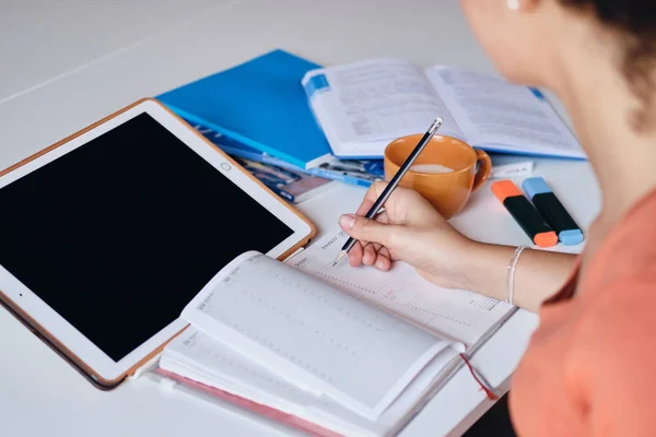 Feche a foto da menina sentada à mesa com tablet e xícara de café escrevendo no caderno estudando em casa — Fotografia de Stock