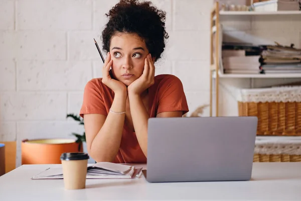 Giovane studentessa pensierosa con i capelli ricci scuri seduta a tavola con laptop e tazza di caffè per andare appoggiata sulle mani pensierosamente guardando da parte studiando a casa accogliente — Foto Stock