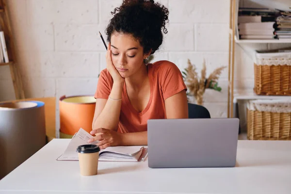 Молодая студентка с темными кудрявыми волосами сидит за столом с ноутбуком и чашкой кофе, чтобы пойти опираясь на руку задумчиво учится в уютном доме — стоковое фото
