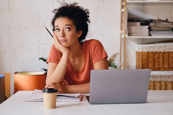 Jovem menina estudante atraente com cabelo encaracolado escuro sentado à mesa com laptop e xícara de café para ir inclinado na mão sonhadoramente olhando para cima estudando na moderna casa aconchegante — Fotografia de Stock