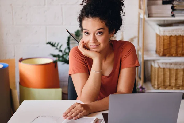 Junge schöne Studentin mit dunklem lockigem Haar sitzt am Tisch mit Laptop auf der Hand gelehnt glücklich in die Kamera schauen Studium an modernen gemütlichen Zuhause — Stockfoto