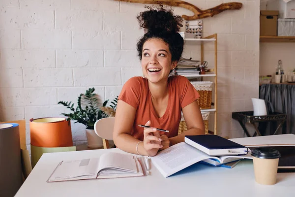 Fiatal vidám tanuló lány, sötét göndör hajú ül az asztalnál, laptop és csésze kávét menni gazdaság ceruzával a kézben boldogan nézett félre tanuló otthonos otthon — Stock Fotó