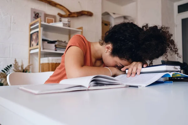年轻的疲惫的学生女孩,黑卷发坐在桌旁,睡在课本上准备考试在舒适的家 — 图库照片