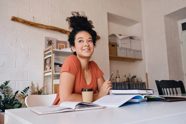 Jovem menina estudante muito sorridente com cabelo encaracolado escuro sentado à mesa com laptop e xícara de café para ir segurando lápis na mão alegremente olhando para o lado estudando em casa aconchegante sozinho — Fotografia de Stock