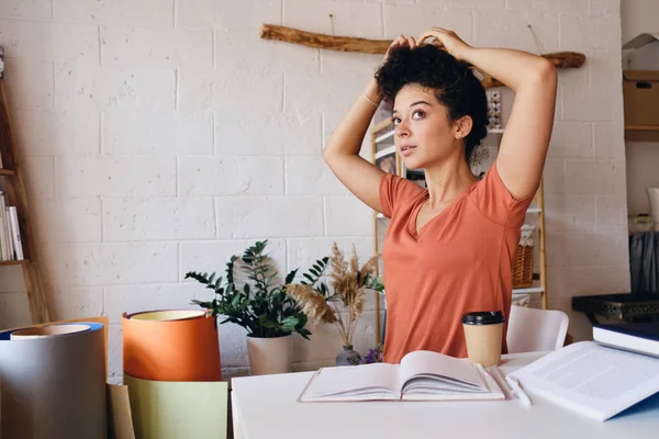 Young pretty student girl with dark curly hair sitting at the table with cup of coffee to go and textbooks thoughtfully looking aside at home — Stock Photo, Image