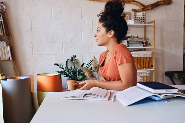 Young attractive student girl with dark curly hair sitting at the table with cup of coffee to go in hand and notebooks around dreamily looking aside studying at home