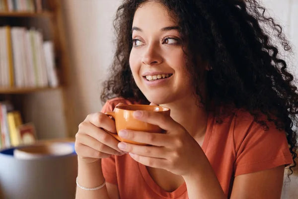 Retrato de jovem atraente mulher sorridente com cabelo encaracolado escuro segurando xícara de café laranja em mãos alegremente olhando de lado em casa aconchegante sozinho — Fotografia de Stock