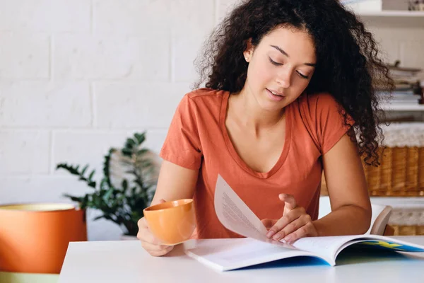 Gadis cantik dengan rambut keriting gelap duduk di meja dengan secangkir kopi oranye di tangan bermimpi membaca buku di rumah yang nyaman — Stok Foto