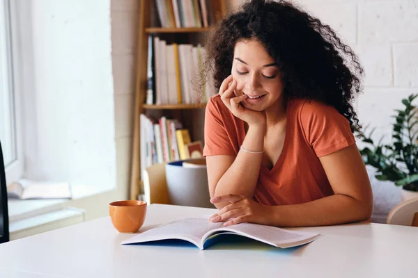 Молодая красивая улыбающаяся женщина с темными вьющимися волосами сидит за столом возле окна с оранжевой чашкой кофе мечтательно читает книгу в современном уютном доме — стоковое фото