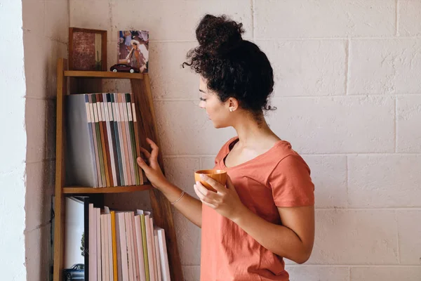 年轻的美丽女人,黑卷发在T恤拿着一杯咖啡在手梦寐以求的选择书从书架在舒适的家 — 图库照片