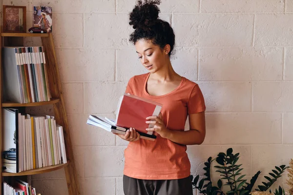 Молодая красивая женщина с темными кудрявыми волосами в футболке, держа книги в руках, стоящие возле книжной полки в уютном доме — стоковое фото