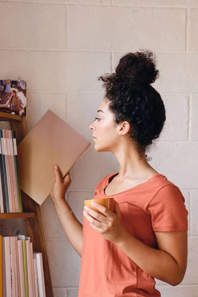 Молода красива жінка з темним кучерявим волоссям у футболці тримає чашку кави в руці, продумано вибираючи книгу з книжкової полиці вдома — стокове фото