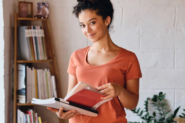 Młoda Ładna uśmiechnięta kobieta z ciemnymi kręcone włosy w T-shirt trzymając książki w rękach szczęśliwie patrząc w aparacie stoi w pobliżu półki w przytulnym domu — Zdjęcie stockowe