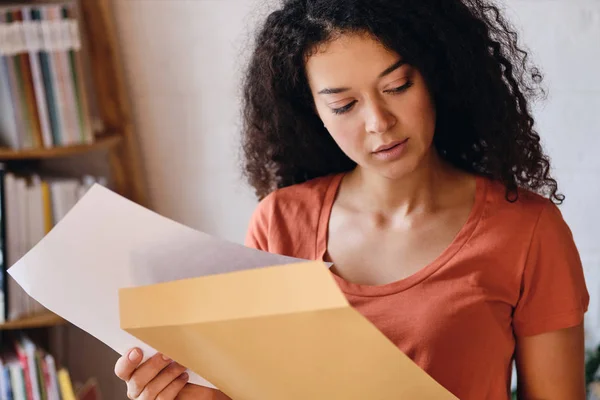 Jonge peinzende Lady met donkere krullend haar in T-shirt zorgvuldig kijken opzij houden brief met examen resultaten in handen thuis — Stockfoto