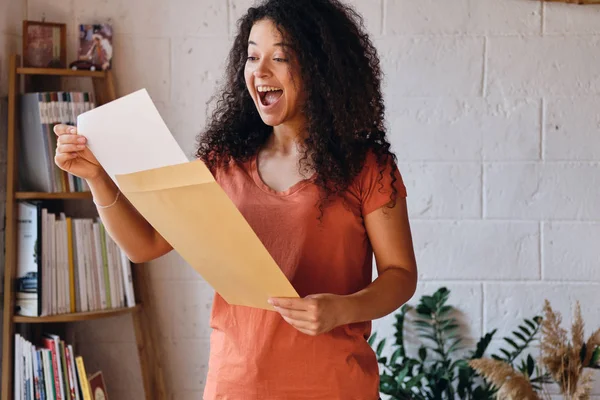 Giovane donna gioiosa con i capelli ricci scuri in T-shirt felicemente apertura busta con risultati dell'esame lettera a casa accogliente — Foto Stock