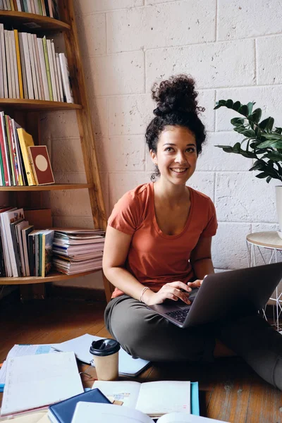 Young beautiful smiling woman with dark curly hair sitting on floor with laptop on knees happily looking aside with books and cup of coffee to go near at cozy home — Stock Photo, Image