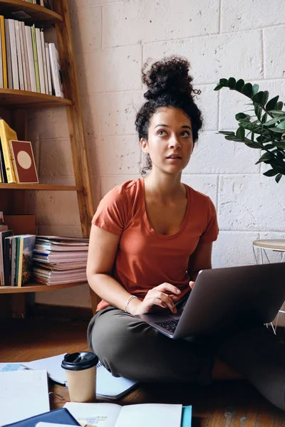 Young attractive woman with dark curly hair sitting on floor with laptop on knees thoughtfully looking aside with books and cup of coffee to go near at cozy home — Stock Photo, Image