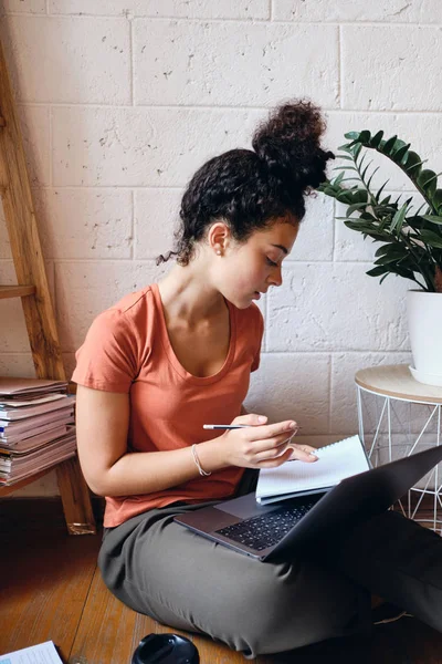 Young student girl with dark curly hair sitting on floor with laptop on knees thoughtfully studying at cozy home alone — Stock Photo, Image