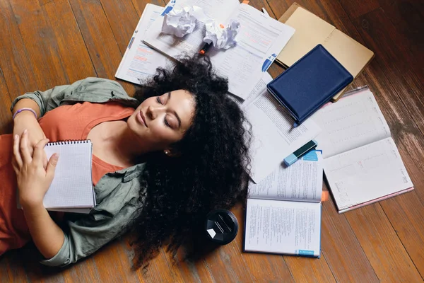 Jovem menina estudante atraente com cabelo encaracolado escuro deitado no chão com livros didáticos ao redor e dormindo em casa aconchegante — Fotografia de Stock