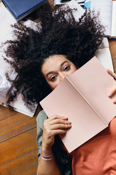 Gadis muda yang menarik dengan rambut keriting gelap tergeletak di lantai dengan buku-buku teks di sekitarnya dengan senang hati melihat ke kamera menutupi mulut dengan buku catatan di rumah — Stok Foto