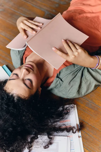 Jovem bela menina estudante sorridente com cabelo encaracolado escuro deitado no chão com livros didáticos em torno de estudar alegremente em casa — Fotografia de Stock