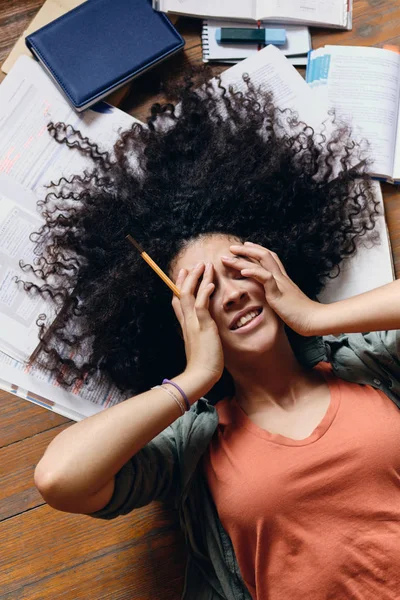 Jovem menina estudante sorridente com cabelo encaracolado escuro deitado no chão com livros didáticos em torno de olhos que cobrem sonhadamente com as mãos em casa — Fotografia de Stock