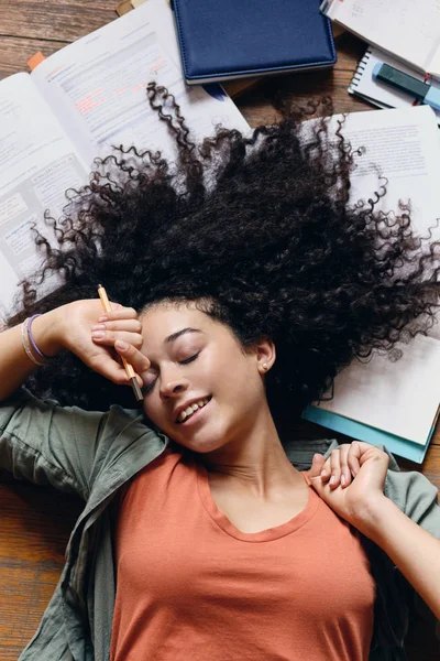 年轻漂亮的微笑学生女孩与黑卷发躺在地板上与教科书周围愉快地睡在家里 — 图库照片