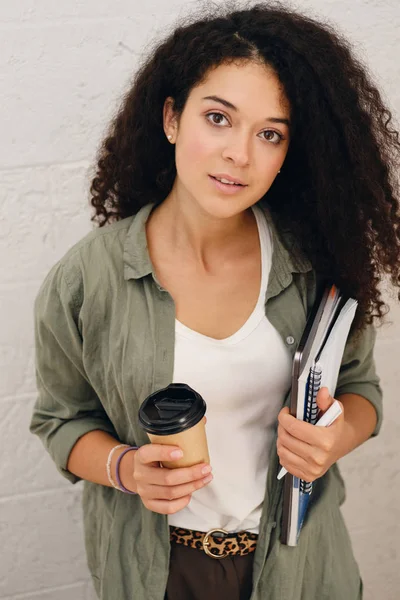 Молодая красивая леди с темными кудрявыми волосами в рубашке цвета хаки держит ноутбук с блокнотом и чашкой кофе, чтобы пойти в руках мечтательно глядя в камеру — стоковое фото