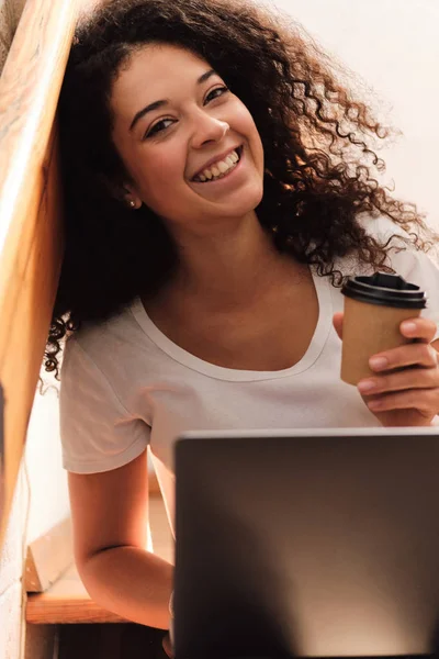 Junge schöne lächelnde Studentin mit dunklem lockigem Haar, die mit Laptop und Tasse Kaffee in der Hand auf der Treppe der Universität sitzt und freudig in die Kamera blickt — Stockfoto