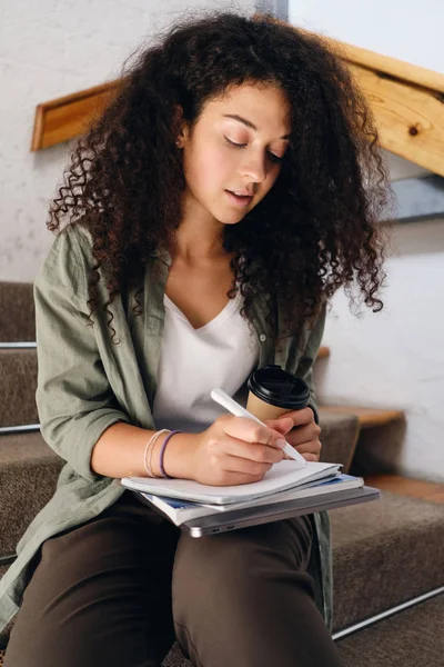 Gadis muda yang menarik dengan rambut keriting gelap duduk di tangga di universitas dengan secangkir kopi untuk pergi di tangan dan bijaksana menulis di notebook — Stok Foto