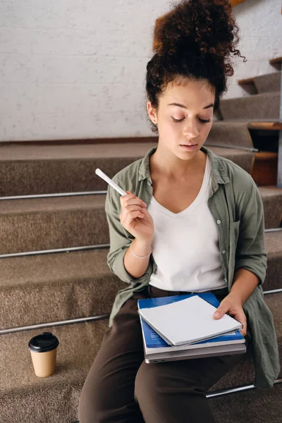Молодая задумчивая студентка с темными кудрявыми волосами сидит на лестнице в университете с чашкой кофе, чтобы подойти вдумчиво задумчиво учится в одиночку — стоковое фото