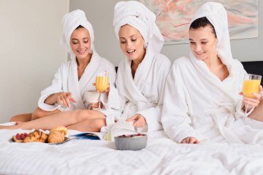 Young pretty smiling women in white bathrobes and towels on head happily reading book together. Attractive girls drinking orange juice having breakfast in bed in modern hotel clipart