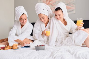 Young beautiful women in white bathrobes and towels on head dreamily reading book together. Pretty girls holding glasses with orange juice in hands having breakfast in bed in modern hotel clipart