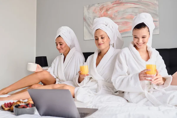 白いバスローブとタオルを頭に乗せた若い美しい笑顔の女性は、一緒にノートパソコンで映画を楽しく見ています。近代的なホテルでベッドで朝食を持つオレンジジュースを飲むかわいい女の子 — ストック写真