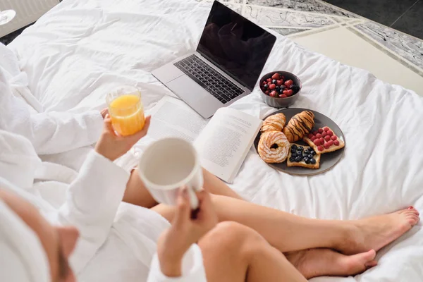 Közelről fotó nők gazdaság pohár gyümölcslé és csésze kávé laptop, könyv és gyönyörű reggeli az ágyban, modern szálloda — Stock Fotó