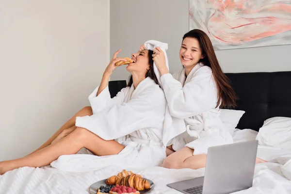 Két gyönyörű nő fehér fürdőköpenyben étkezési péksütemény boldogan időtöltést együtt reggelizik az ágyban a modern szállodában — Stock Fotó