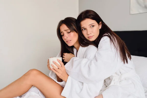 Jonge boos vrouw in witte badjas houden kopje koffie in de hand en huilen terwijl peinzende meisje zorgvuldig knuffelen en haar steunen op groot bed thuis — Stockfoto