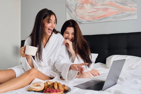 Две веселые женщины в белых халатах счастливо смотрят фильм на ноутбуке вместе с вкусной выпечкой и кофе. Улыбающиеся девушки завтракают в постели в современном отеле — стоковое фото