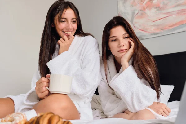 Jovem bela mulher sorridente com xícara de café na mão feliz assistindo filme enquanto menina bonita perto pensativo olhando de lado. Duas meninas atraentes tomando café da manhã na cama em casa — Fotografia de Stock