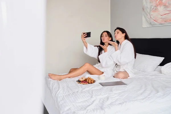 Zwei attraktive Frauen in weißen Bademänteln, die verträumt Luftküsse versenden und zusammen mit dem Handy Fotos machen. schöne Mädchen frühstücken im Bett mit Laptop im modernen Hotel — Stockfoto