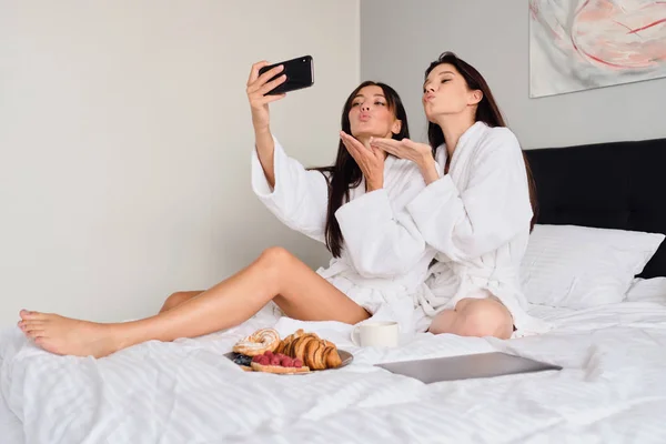 Duas mulheres bonitas em roupões de banho brancos alegremente enviando beijos aéreos tirando fotos juntas no celular. Meninas bonitas tomando café da manhã na cama com laptop no hotel moderno — Fotografia de Stock