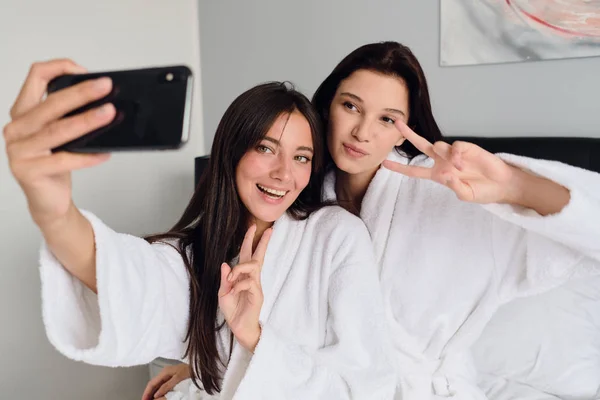 Jonge mooie glimlachende vrouwen in witte badjassen die gelukkig twee vingers laten zien en foto's samen nemen op mobiele telefoon bij Cozy Home — Stockfoto