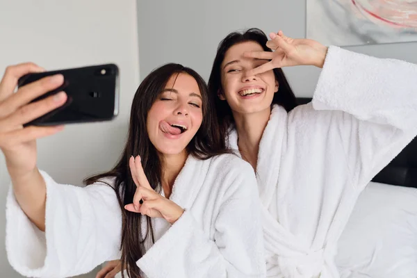 Jonge mooie glimlachende vrouwen in witte badjassen die gelukkig twee vingers gebaar laten zien en coole Foto's samen nemen op mobiele telefoon bij Cozy Home — Stockfoto