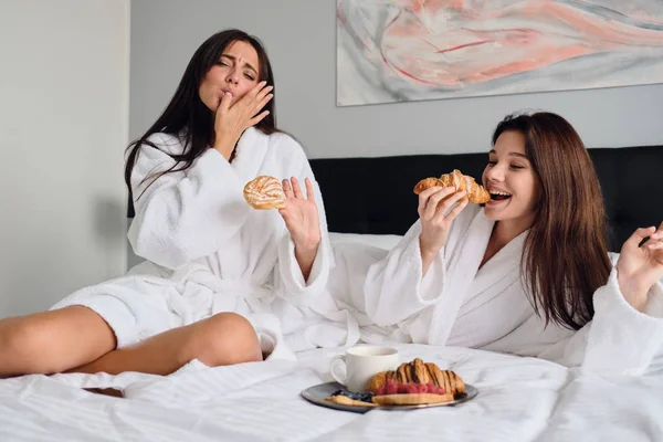 Duas jovens mulheres bonitas em roupões de banho brancos comendo deliciosa pastelaria juntos. Meninas atraentes tomando café da manhã na cama no moderno hotel acolhedor — Fotografia de Stock